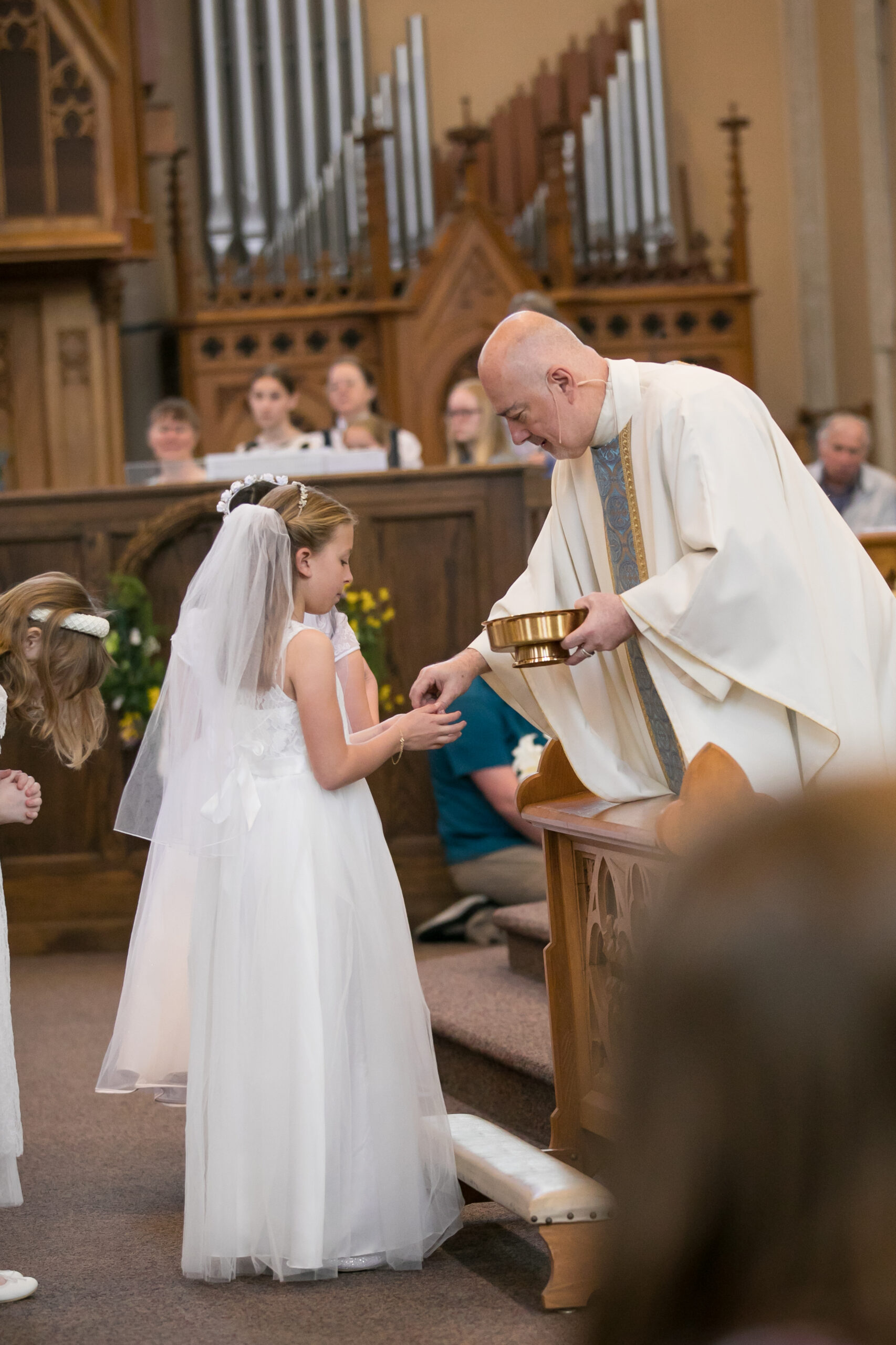 A girl in her First Communion dress receiving the Eucharist from the priest
