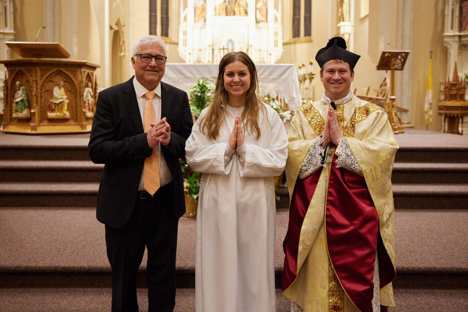A young woman in a white robe with her sponsor and a priest at the altar