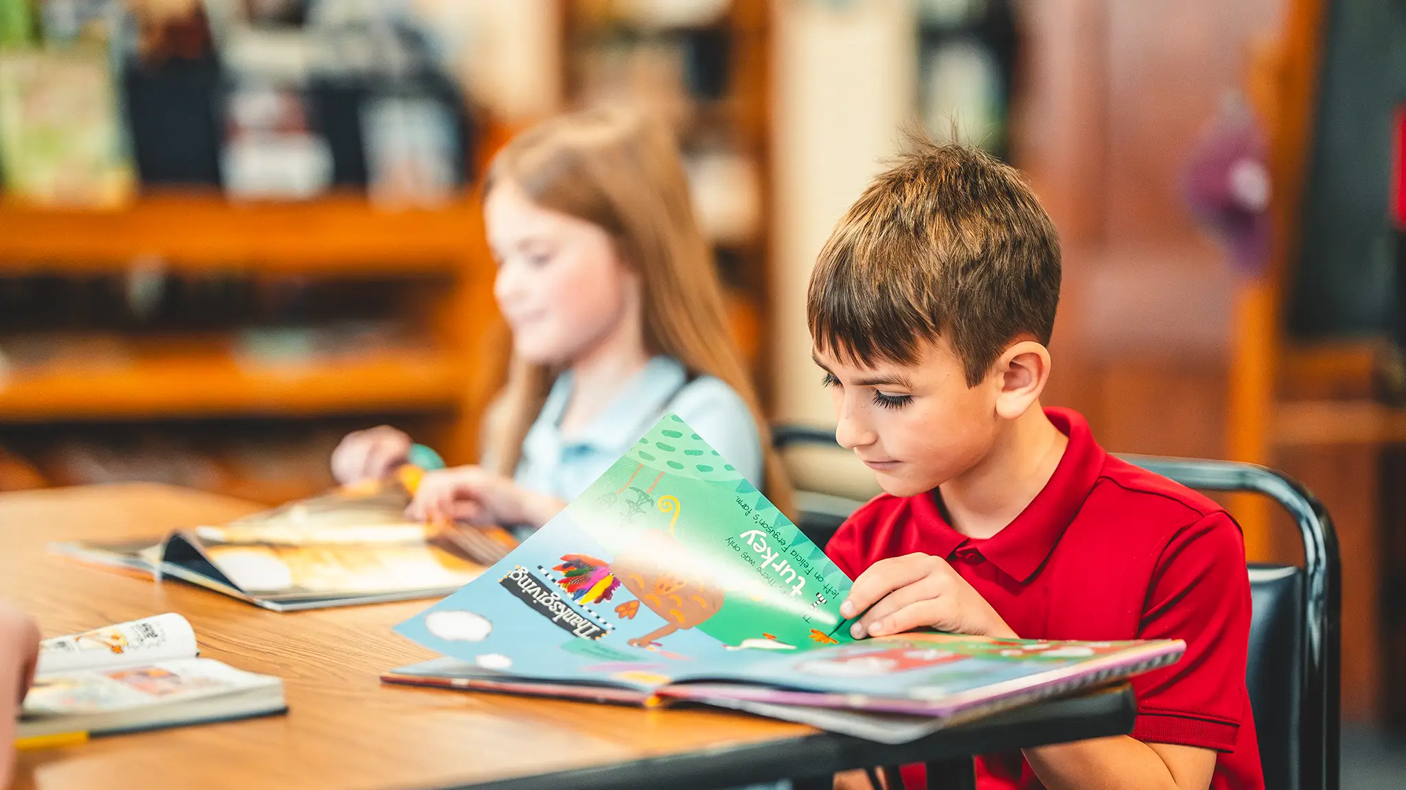 A St. Francis boy in a red uniform shirt reading a picture book at a library table