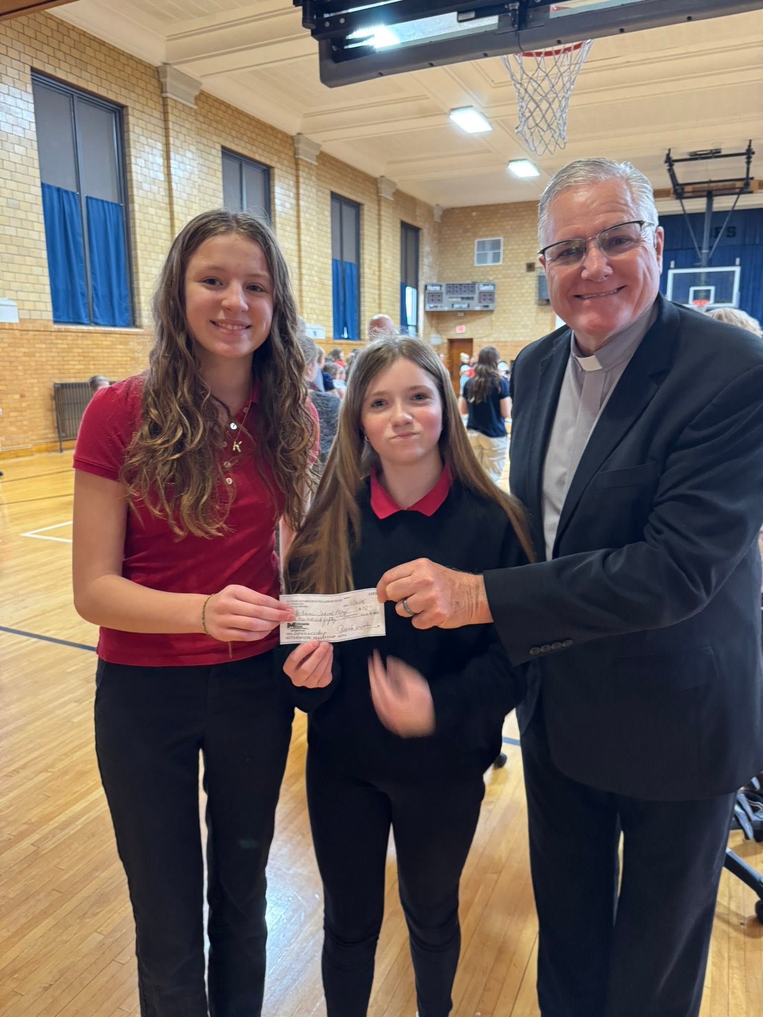 Two St. Francis students receiving a scholarship check from a priest in the school gym