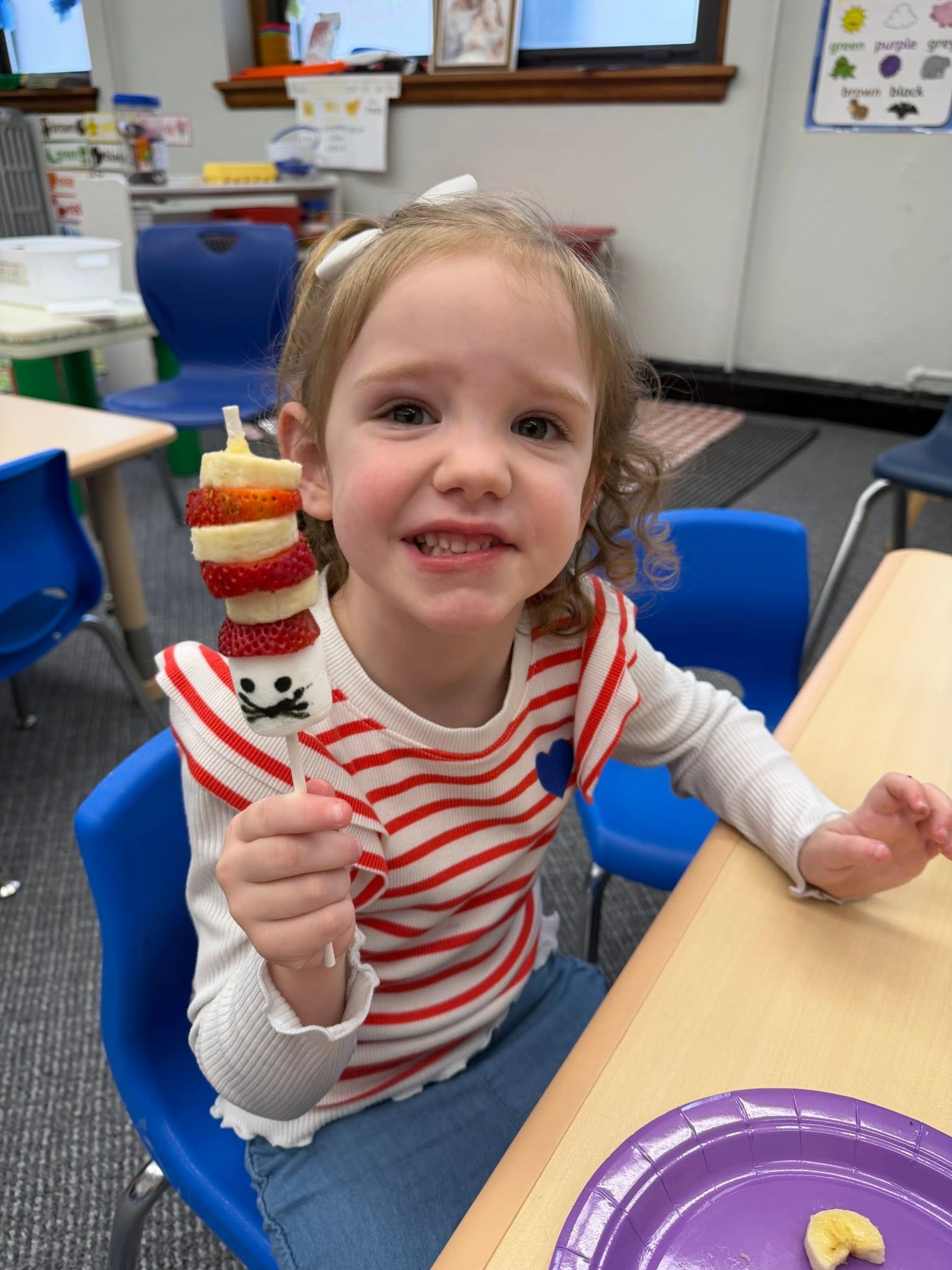 A smiling St. Francis preschooler holding up a fruit kabob she made in class