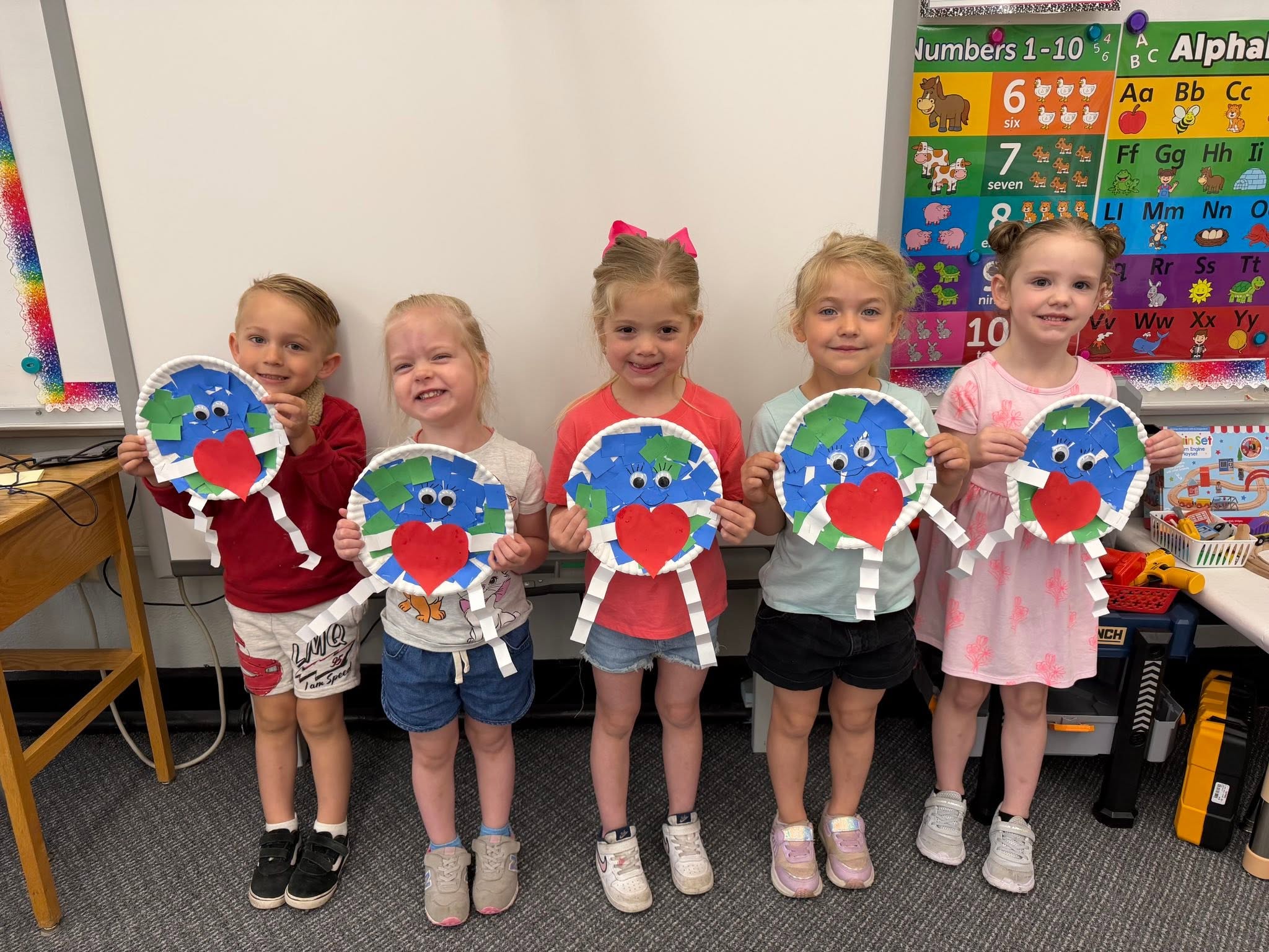 Five St. Francis preschoolers holding paper plate Earth crafts in front of the classroom alphabet chart