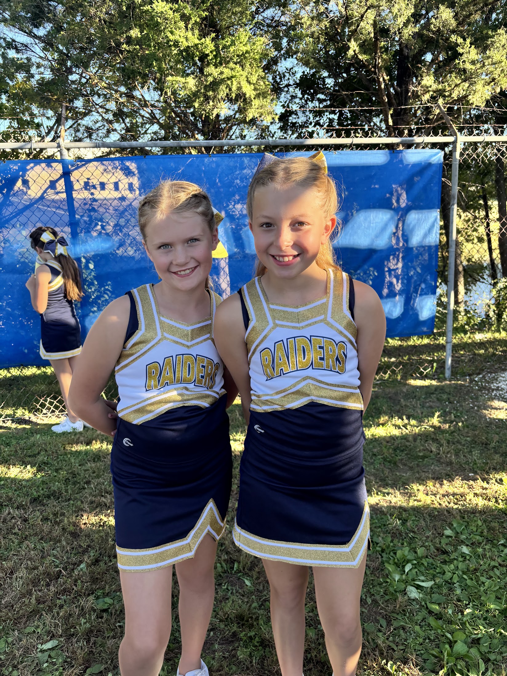 Two young Raiders cheerleaders in navy and gold uniforms smiling outside at a game