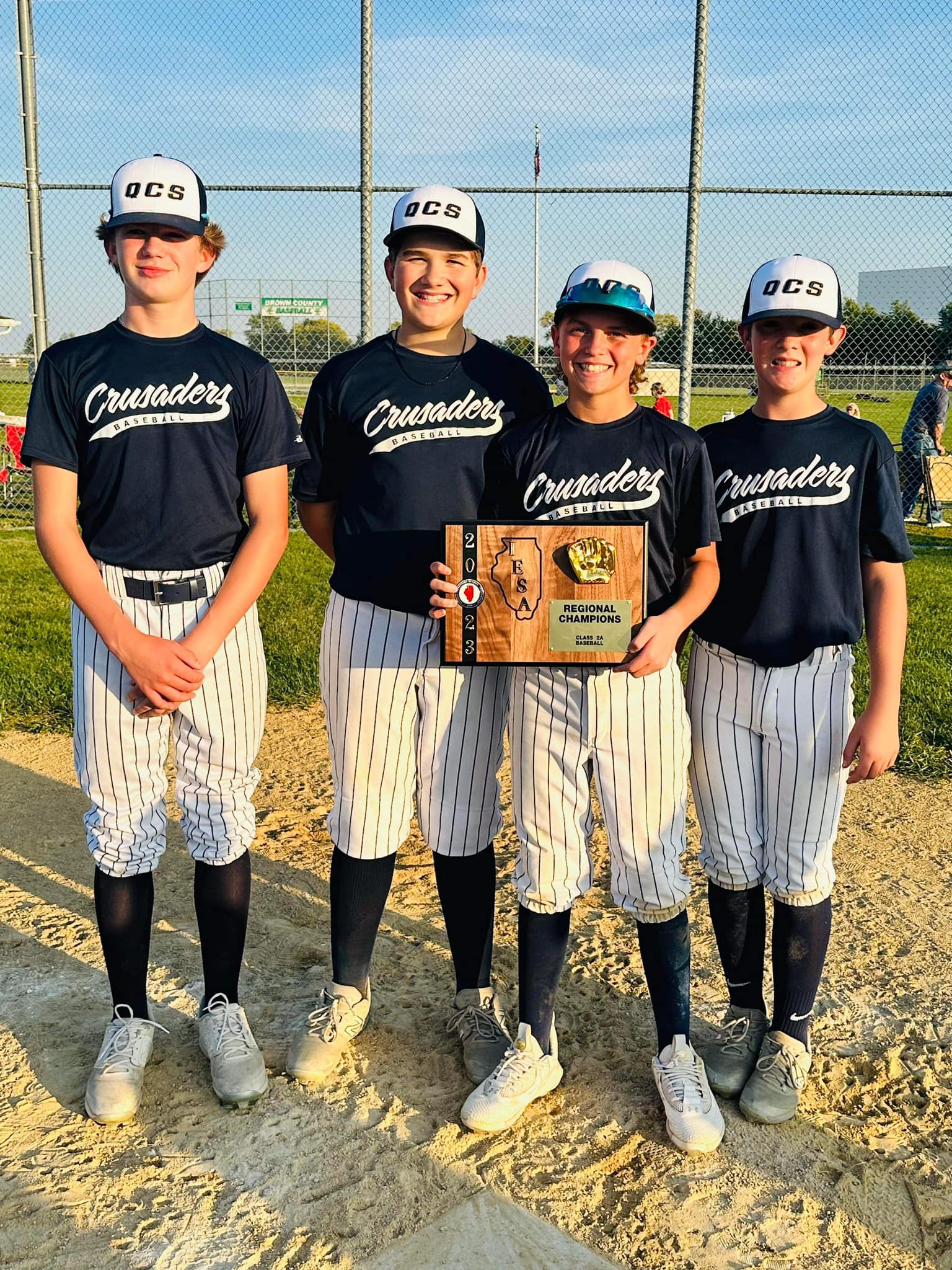 Four Quincy Catholic Crusaders baseball players in pinstripes holding a championship plaque