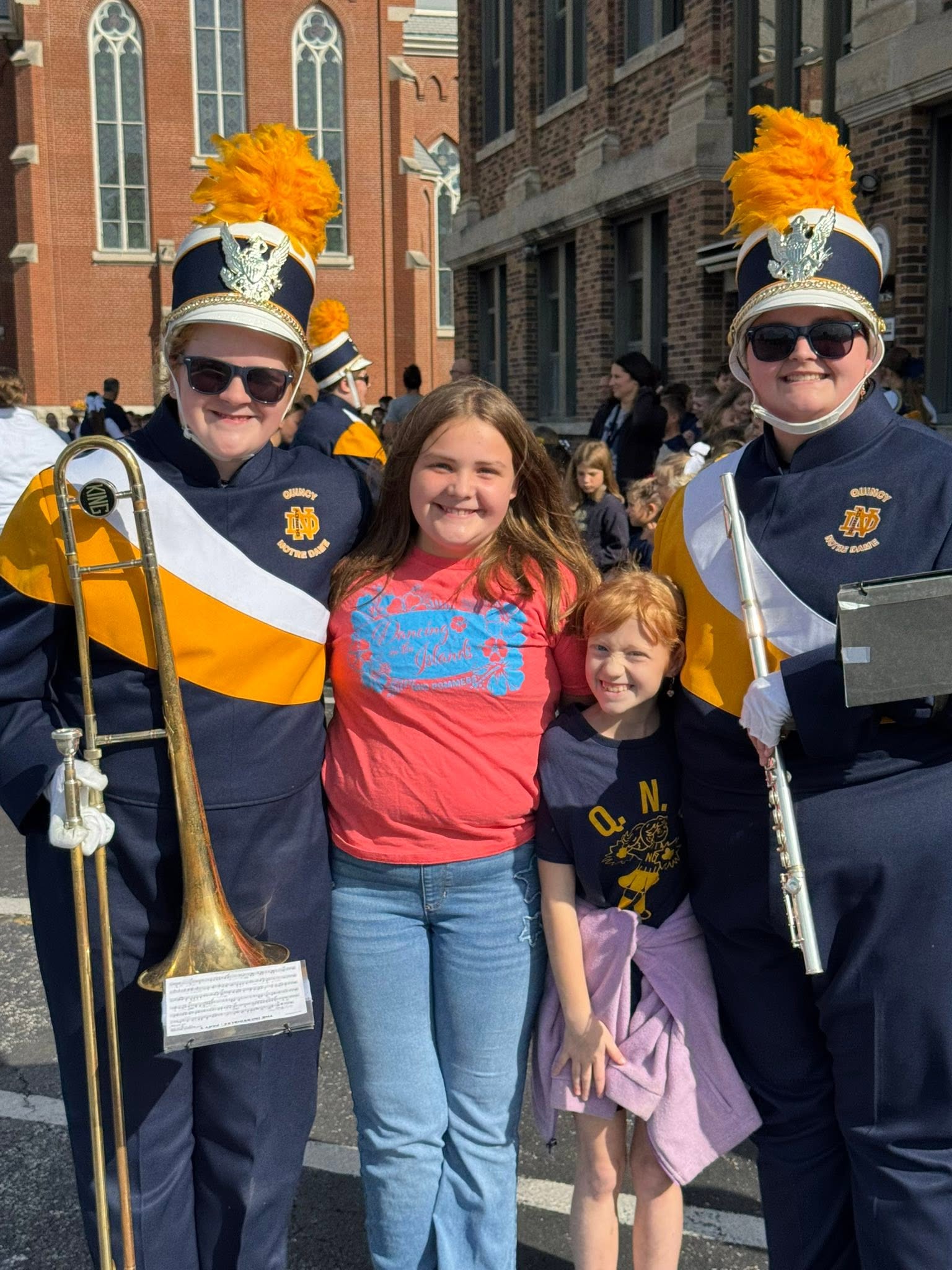 Two Quincy Notre Dame Raiders marching band members in plumed uniforms, one with a trombone, standing with two younger girls