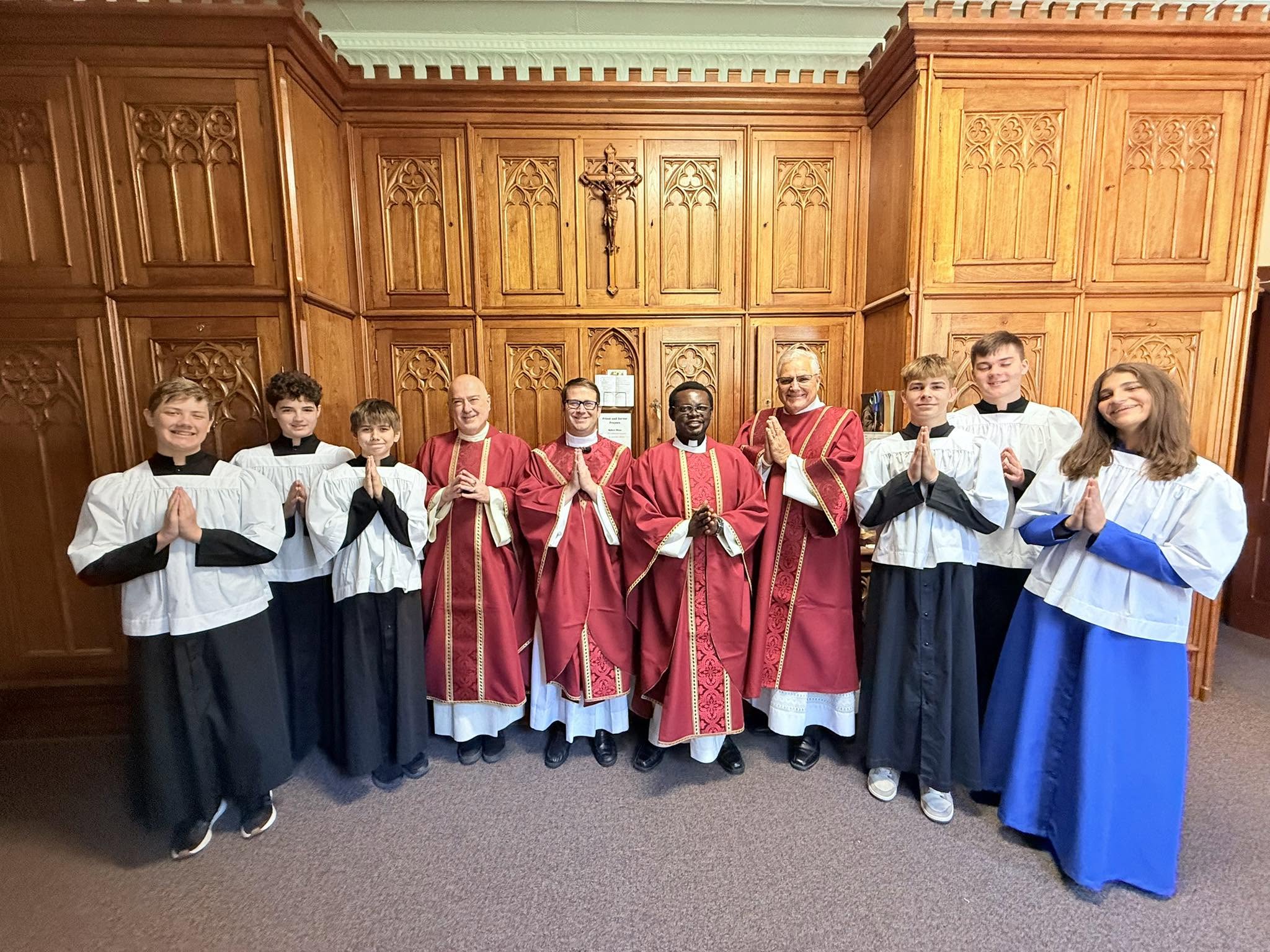 St. Francis altar servers in cassocks and surplices standing with priests in red vestments in the sacristy