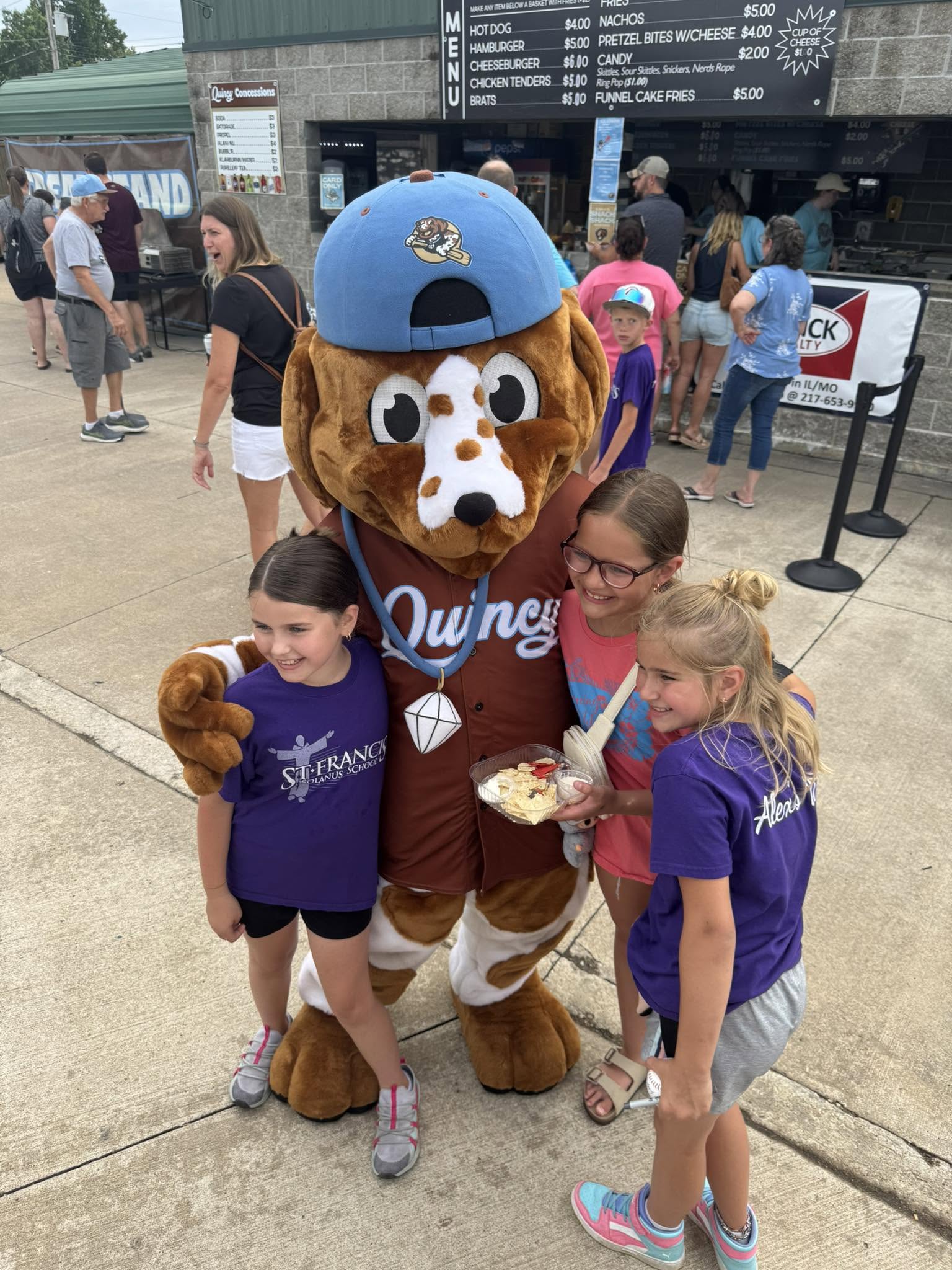 St. Francis girls in purple school shirts hugging the Quincy Doggy Paddlers mascot at a ballgame