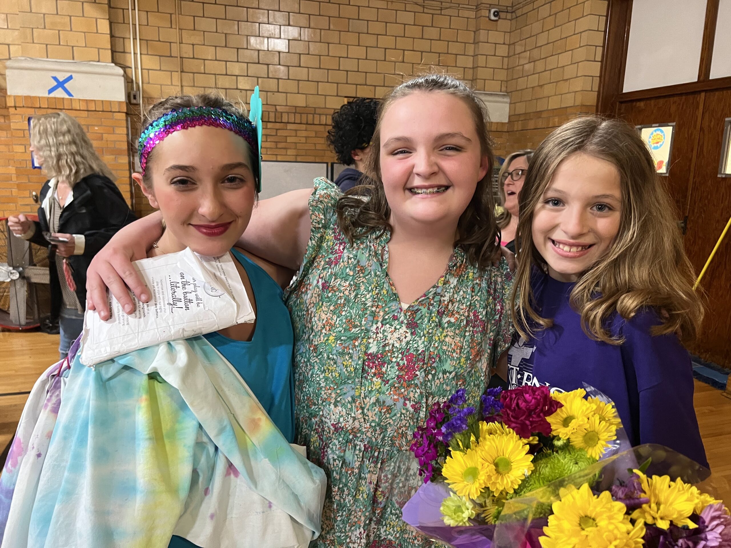 Three smiling St. Francis girls after a school play, one holding a script and another a bouquet of sunflowers